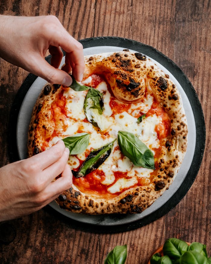 pexels photo 10068752 Close-up of hands adding basil to a freshly baked Neapolitan pizza, highlighting its cheesy and delicious appeal.
